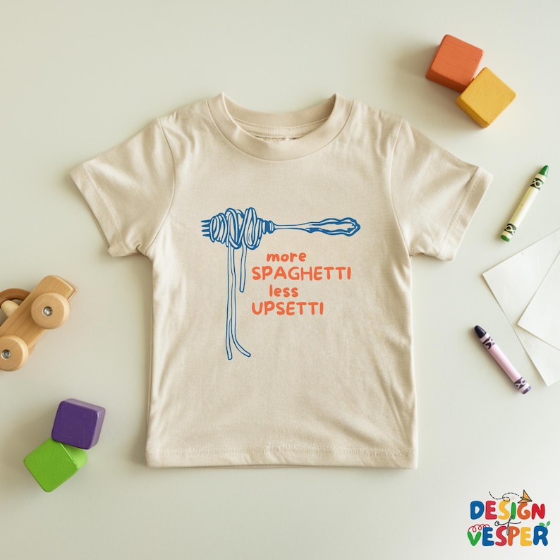 Pu&ograve; includere: Una maglietta beige con la scritta "more spaghetti less upsetti" in blu e arancione. La grafica mostra una forchetta con spaghetti. La maglietta &egrave; circondata da giocattoli e pastelli colorati.
