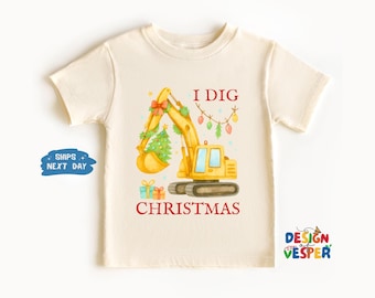 以下が含まれることがあります： クリーム色のTシャツ。クリスマスツリーとライトで飾られた黄色の掘削機が描かれています。「I DIG CHRISTMAS」の文字が赤色でプリントされています。ホリデーシーズン向けのお祝いのデザインです。