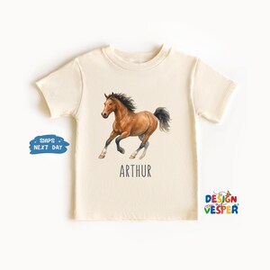 May include: Cream-colored t-shirt featuring a watercolor illustration of a brown horse in motion. The horse has a black mane and tail, with the name "ARTHUR" printed below. The shirt is a children's apparel item.