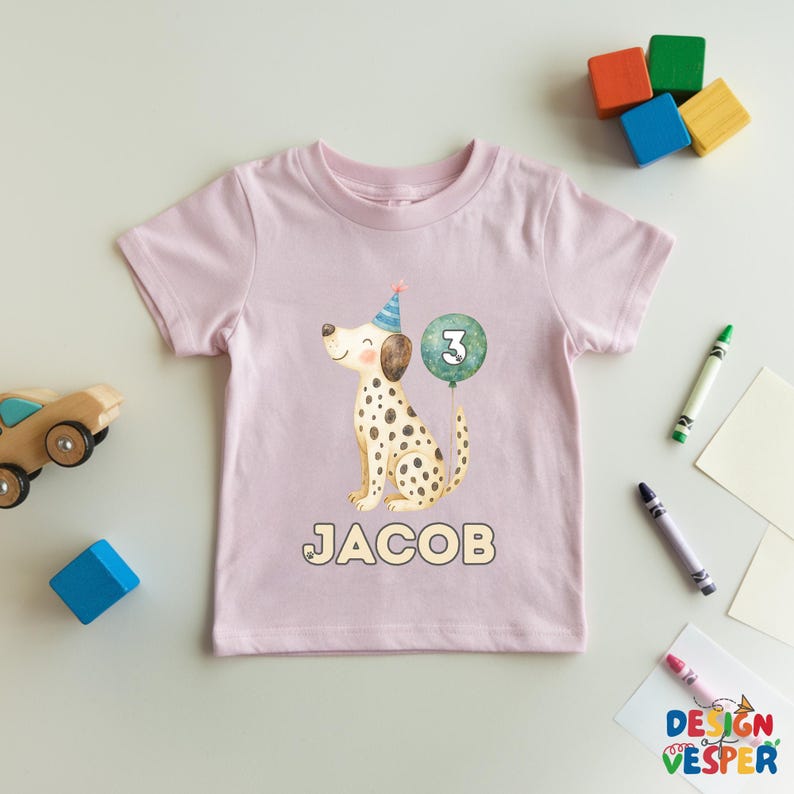 May include: A light pink toddler t-shirt featuring a Dalmatian dog illustration wearing a party hat and holding a number 3 balloon. The name "JACOB" is printed below the dog. The shirt is surrounded by colorful toys and crayons.