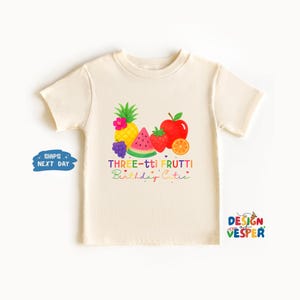 May include: Cream-colored toddler t-shirt featuring a colorful fruit graphic. The design includes a pineapple, watermelon, strawberry, apple, and orange. Text reads "THREE-tti FRUTTI Birthday Cutie" in rainbow colors.