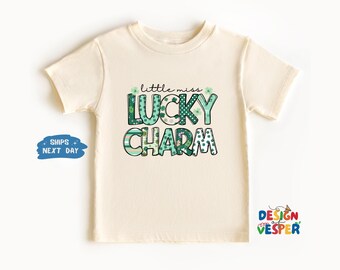 以下が含まれることがあります： クリーム色のTシャツ。緑と白で「little miss LUCKY CHARM」の文字がプリントされています。デザインにはクローバーと装飾的なフォントが含まれています。子供服です。