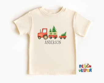 May include: Cream-colored t-shirt featuring a watercolor illustration of a red tractor pulling two wagons filled with Christmas trees. The name "ANDERSON" is printed below the image. The shirt has short sleeves and a crew neck.