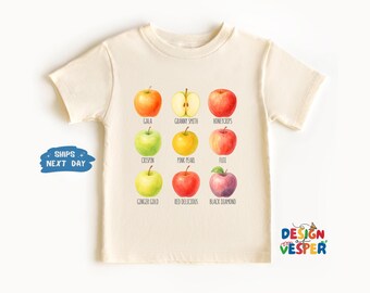 May include: Cream-colored t-shirt featuring a watercolor illustration of nine different apple varieties, each labeled with its name. The apples are in shades of red, green, and yellow. The shirt also has the text "DESIGN VESPER".