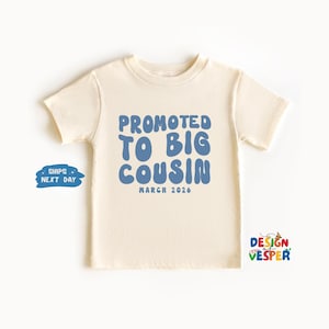 May include: A cream-colored toddler t-shirt with the text "PROMOTED TO BIG COUSIN" in blue bubble letters. Below the text is "MARCH 2026" in a smaller font. The shirt has short sleeves and a crew neck.