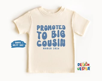 以下が含まれることがあります： クリーム色の幼児用Tシャツ。青い泡文字で「PROMOTED TO BIG COUSIN」と書かれています。その下に小さなフォントで「MARCH 2026」と書かれています。このシャツは半袖で、クルーネックです。