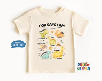 以下が含まれることがあります： 「GOD SAYS I AM」の文字とカラフルな恐竜のイラストが描かれたクリーム色のTシャツ。「ハンサム」「親切」「強い」などの言葉と聖書からの引用が含まれています。Design Vesperによるデザイン。