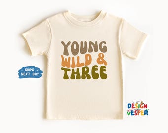 May include: A cream colored t-shirt with the text "YOUNG WILD & THREE" in brown, orange, and green letters. The letters are in a retro font.