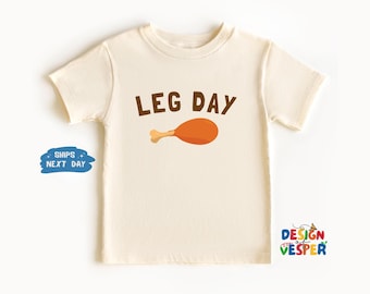 以下が含まれることがあります： 茶色の文字で「LEG DAY」と書かれた、鶏もも肉のイラストが描かれたクリーム色の子供用Tシャツ。Tシャツは半袖で丸首です。背景は白です。