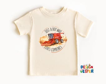 以下が含まれることがあります： クリーム色の幼児用Tシャツ。赤いコンバインハーベスターが畑にいる円形のグラフィックが特徴です。「JUST A BOY WHO LOVES COMBINES」の文字が画像の周りにプリントされています。シャツは半袖で、クルーネックです。