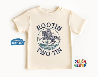 May include: Cream-colored t-shirt featuring a blue graphic of a cowboy riding a horse, with the words "ROOTIN TWO-TIN" in a circular design. The shirt is a short-sleeved style, suitable for children's wear.
