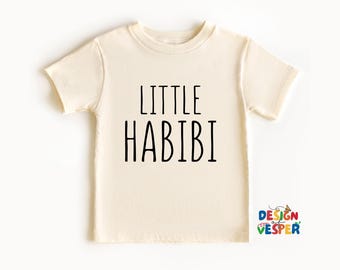 以下が含まれることがあります： 「LITTLE HABIBI」の黒い文字がプリントされた、クリーム色の半袖Tシャツ。クルーネックで、右下に小さなロゴが入っています。