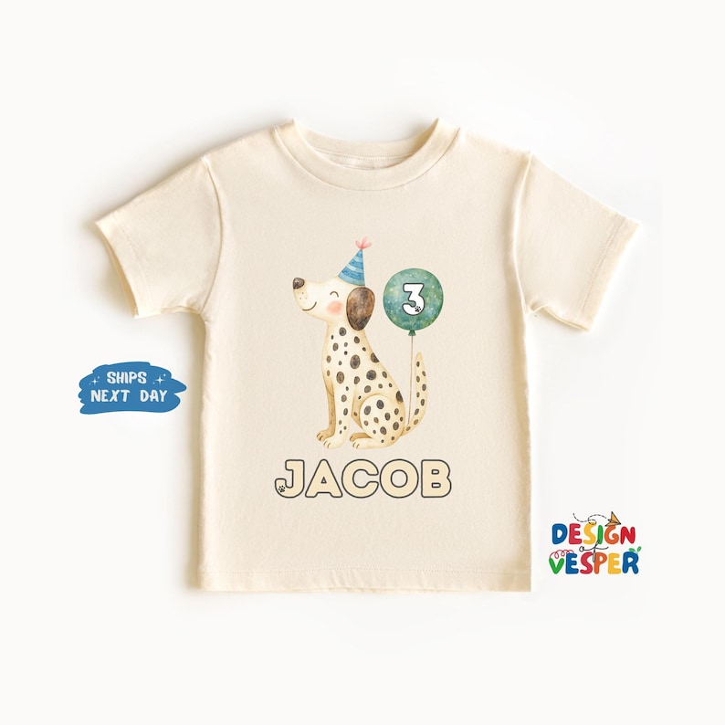 May include: Cream-colored toddler t-shirt featuring a watercolor illustration of a Dalmatian dog wearing a party hat and holding a number 3 balloon. The name "JACOB" is printed below the dog in a bold font. The shirt is a short-sleeved design.