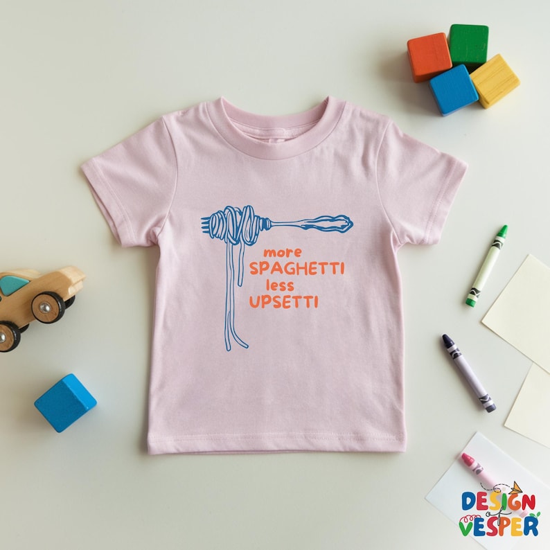 Pu&ograve; includere: Maglietta rosa chiaro per bambini con un'illustrazione blu di spaghetti su una forchetta. Il testo arancione "more spaghetti less upsetti" &egrave; stampato. Giocattoli e pastelli colorati circondano la maglietta.