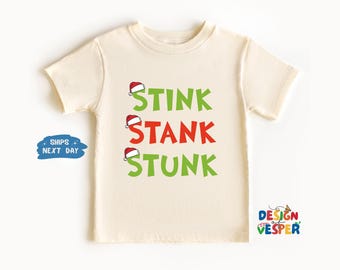以下が含まれることがあります： クリーム色の幼児用Tシャツ。緑と赤で「STINK STANK STUNK」の文字がプリントされており、各単語の上にはサンタ帽が描かれています。Tシャツは半袖で、クルーネックです。デザインは前面の中央に配置されています。
