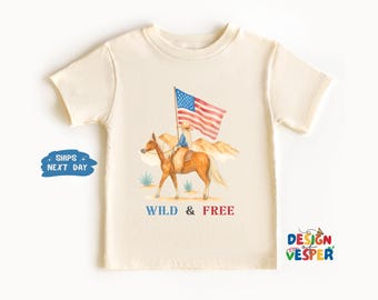以下が含まれることがあります： クリーム色のTシャツ。水彩画で、アメリカの国旗を持った人が馬に乗っているイラストが描かれています。「WILD & FREE」の文字が下にあります。半袖でクルーネックのシャツです。