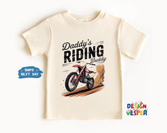 以下が含まれることがあります： クリーム色のTシャツ。モトクロスバイクのイラストと「Daddy's Riding Buddy」の文字がプリントされています。グラフィックは、赤と黒のダートバイクが土煙を上げている様子です。Tシャツは半袖で、クルーネックです。