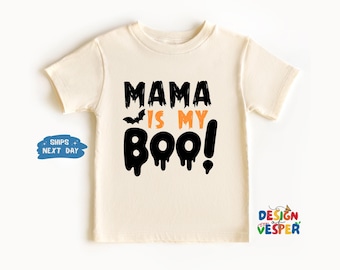 以下が含まれることがあります： クリーム色の子供用Tシャツ。黒とオレンジで「MAMA IS MY BOO！」と書かれています。「BOO！」という言葉の上には、小さな黒いコウモリのグラフィックがあります。このシャツは半袖のクルーネックです。
