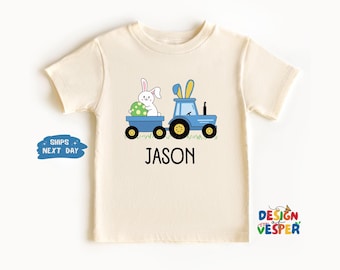 以下が含まれることがあります： 薄いベージュのTシャツに、青いトラクターと緑のイースターエッグを持った白いウサギが描かれています。トラクターの下には「JASON」という文字が印刷されています。