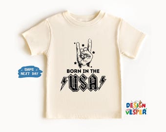 以下が含まれることがあります： クリーム色のTシャツ。黒い手のジェスチャーと「BORN IN THE USA」の文字がプリントされています。デザインには星と稲妻のアクセントがあり、ロックンロールをテーマにしています。子供向けです。