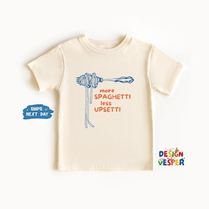 Pu&ograve; includere: T-shirt color crema con grafica blu di spaghetti e forchetta. Il testo "more spaghetti less upsetti" &egrave; in arancione e blu. La maglietta &egrave; a maniche corte e girocollo.