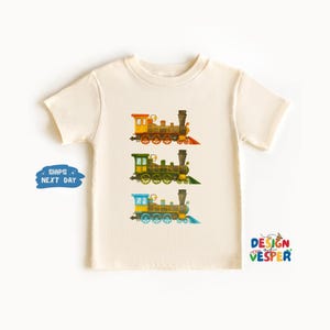 May include: Cream-colored t-shirt featuring three colorful train illustrations. The trains are in shades of yellow, green, and blue, each with black smokestacks and red, orange, and blue accents. The shirt has the text "DESIGN VESPER".