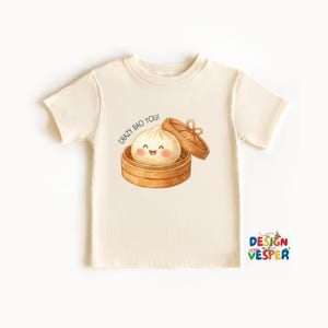 May include: Cream-colored t-shirt featuring a watercolor illustration of a smiling bao bun in a bamboo steamer. The text "CRAZY BAO YOU!" is above the bao bun. The shirt has short sleeves and a crew neck. The Design Vesper logo is in the bottom right corner.