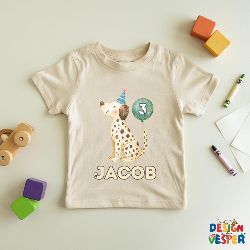 May include: A beige toddler t-shirt featuring a watercolor illustration of a Dalmatian dog wearing a party hat and holding a balloon with the number 3. The name "JACOB" is printed below the dog. The shirt is surrounded by children's toys.
