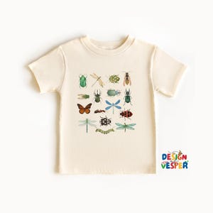 May include: Cream-colored t-shirt featuring a collection of watercolor insects, including butterflies, dragonflies, and beetles. The insects are in various colors, such as green, orange, and blue. The shirt has short sleeves and a round neckline.