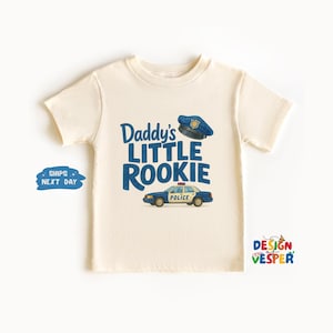 May include: Cream-colored t-shirt with the text "Daddy's Little Rookie" in blue, along with a police hat and car graphic. The shirt has short sleeves and a crew neck. A blue and white graphic that says "Ships Next Day" is on the left.