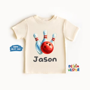 May include: Cream-colored t-shirt featuring a watercolor illustration of bowling pins and a red bowling ball. The name "Jason" is printed in gray below the graphic. The shirt has short sleeves and a crew neck.