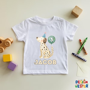 May include: White toddler t-shirt featuring a watercolor illustration of a Dalmatian dog wearing a party hat and holding a balloon with the number 3. The name "JACOB" is printed below the dog. The shirt is decorated with a design from Design Vesper.