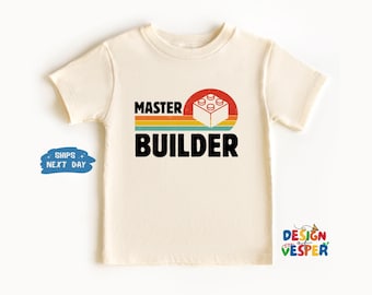 May include: Cream-colored t-shirt with the words "MASTER BUILDER" in black block letters. A red and orange circle with a white Lego block design is above the word "BUILDER". The shirt is a short-sleeved crew neck.