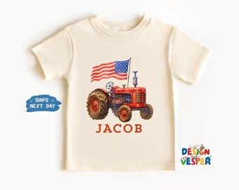 以下が含まれることがあります： クリーム色のTシャツ。赤いトラクター、アメリカ国旗、赤いブロック体の名前「JACOB」が描かれています。トラクターは、大きくて濃い茶色のタイヤが付いています。子供服です。