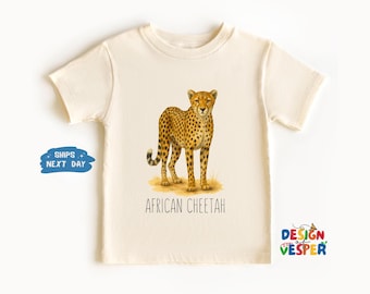 May include: Cream-colored t-shirt featuring a detailed illustration of an African cheetah. The cheetah is depicted with its distinctive spotted coat and is standing on a patch of grass. The words "AFRICAN CHEETAH" are printed below the image.