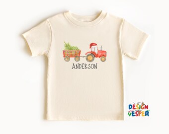 May include: Cream-colored t-shirt featuring a watercolor illustration of a red tractor pulling a wagon filled with a Christmas tree. The name "ANDERSON" is printed below the tractor. The shirt has short sleeves and a crew neck.