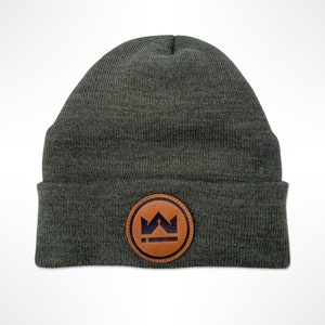 May include: Olive green beanie with a brown leather patch. The patch features a crown and cross design within a circular border. The beanie is made of a knitted material and is suitable for cold weather.