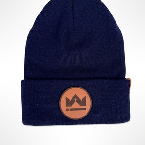 Peut inclure: Un bonnet bleu marine avec un écusson en cuir marron. L'écusson présente un motif de couronne et de croix. Le bonnet est en maille et possède un bord replié. Un accessoire d'hiver élégant.