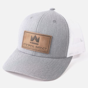 May include: A gray trucker hat with a white mesh back and a curved brim. The hat features a brown leather patch with the text "ETERNAL IMPACT KINGDOM FOCUSED APPAREL" and a crown logo.