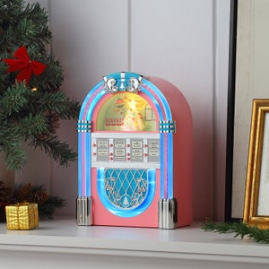 Könnte beinhalten: Eine rosafarbene und blaue Jukebox im Vintage-Stil mit einem Weihnachtsbaum-Design. Die Jukebox hat ein beleuchtetes Fenster und eine silberne Verzierung. Sie spielt Weihnachtsmusik.