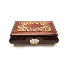 May include: A wooden music box with a clock face on the front. The box is decorated with a gold inlay design of swirling leaves and vines. The box is brown with a reddish hue.