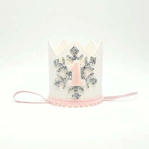 May include: White felt crown with a silver glitter snowflake and a pink felt number one. The crown has a pink elastic band.