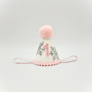 May include: White and pink birthday party hat with a pink pom-pom on top. The hat has a silver glitter snowflake design and the number "1" in pink glitter.