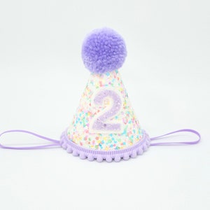 Pode incluir: Chapéu de festa em forma de cone, decorado com confetes coloridos e um grande número "2" com glitter. O chapéu apresenta um pompom lavanda no topo e uma borda lavanda com uma tira de queixo a condizer.