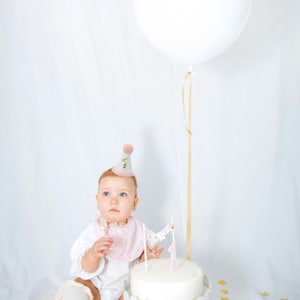 May include: A baby wearing a white dress and pink bib, a white knitted hat with a pink pom-pom and the word "One" embroidered on it, and white knitted leg warmers. The baby is sitting in front of a white cake with pink flags and a white cake stand. A white balloon is in the background.