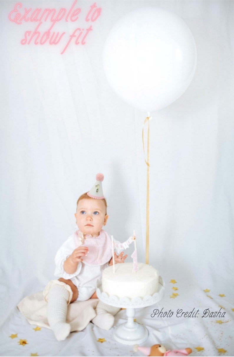 May include: A baby wearing a white dress and pink bib, a white knitted hat with a pink pom-pom and the word "One" embroidered on it, and white knitted leg warmers. The baby is sitting in front of a white cake with pink flags and a white cake stand. A white balloon is in the background.