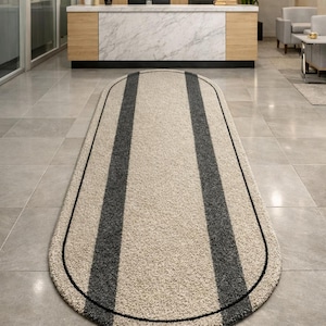 May include: Oval-shaped beige rug with two dark gray vertical stripes and a black border. The rug is placed on a tiled floor in a reception area, adding a touch of style to the space.