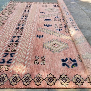 May include: A rolled-up, rectangular rug with a pink and cream background. The rug features geometric patterns in navy blue, black, and cream. The design includes diamond, zigzag, and square shapes. The rug is partially unrolled, revealing the rolled-up section.