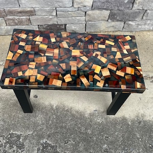 May include: A black coffee table with a unique epoxy resin top. The top is made of many small pieces of wood, creating a mosaic-like pattern. The table has a modern design and would be a great addition to any home.
