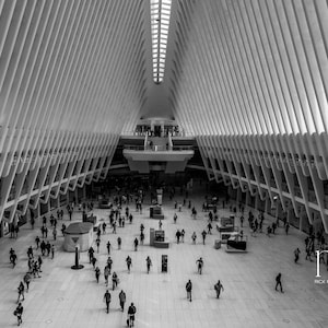 May include: Black and white photograph of a large, modern interior space with a high, vaulted ceiling. The architecture features repeating, angled beams. Numerous people are walking on the polished floor. The image is in black and white.